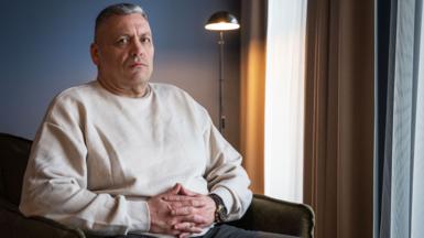 A man with short grey hair and a cream coloured sweat shirt looks into the camera - he is seated on a green armchair with blue and beige curtains behind him