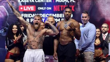 Jake Paul points his arm in the air as he and Anthony Joshua stand side by side at a weigh-in