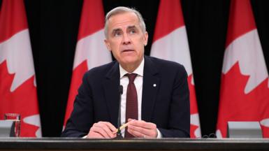 Canada's PM Mark Carney looks to the right of frame while sitting at a press conference briefing table in front of four Canadian flags in Ottowa in May. He wears a black suit and red tie, and holds a pen in his hands with a glass of water beside him on the table.