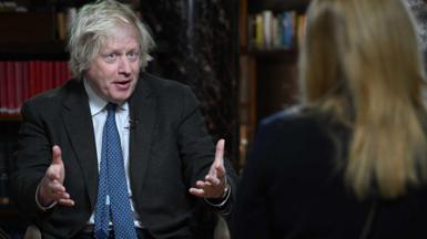 Former prime minister Boris Johnson speaking while holding out both hands. In the foreground, out of focus, we see the back of a woman with blonde hair.