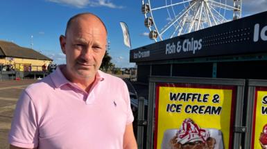 A man in a pink polo shirt looks at the camera. He is standing next to signs advertising waffle and ice cream. A big wheel is in the distance. 