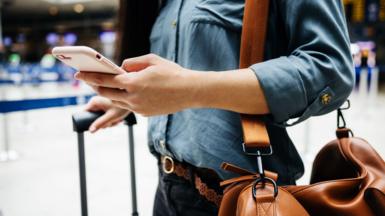 A close up of a woman holding a phone in one hand and a wheelie bag handle in the other hand