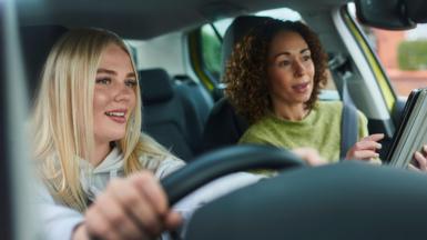 A young blonde woman wearing a white hooded jumper appears to be taking a driving lesson, and is taking instruction from an older, but still quite young woman, who has dark curly hair and a woolen green jumper. They both look like they are looking at something in front of the car 