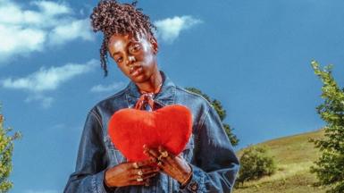 Cat Burns holds up a cushion in the shape of a heart, while standing against a countryside landscape, in a promotional photo for her new album How To Be Human