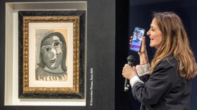 A blue painting by Picasso, in his trademark cubist style, framed on the wall beside a woman holding a microphone up to her mobile phone, which shows a video call with a man wearing glasses - both are smiling broadly. 