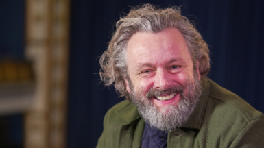 Michael Sheen, who has long grey hair and a beard, smiles to camera while sitting in a theatre 