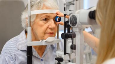 An older lady having an eye test. She is looking into a machine while  an optician hold a lens to her left eye.