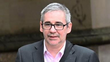 Iain Robertson wears a grey suit and pink shirt. he has short grey hair and wears dark glasses. He is walking outside a building