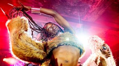 A performer is dramatically lit on-stage, wearing a fur-effect tank top with long, thick sleeves. They are holding their dreadlocks above their head as they dance, wearing jewellery on their wrist and across their face. A second performer is visible behind them with a large, white wig on their head and dramatic eye make-up.