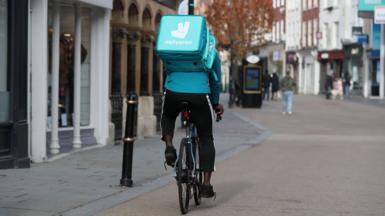 Deliveroo rider on bike wearing black trousers, bright blue top, and bright blue square Deliveroo bag on his back saying the firm's name and with its rabbit logo on it