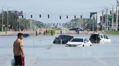 A young man wades through flood waters that come up to the top of car wheels