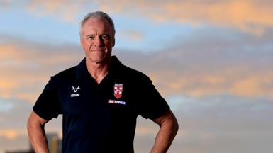 Brian McDermott, wearing a black England polo shirt, looks to the camera, with the sky in the background