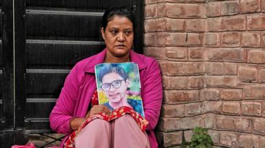 A woman sits holding a picture of her loved one. She is dressed in a bright pink top and pale pink trousers