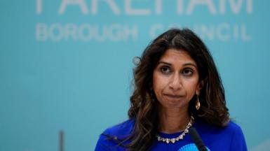 Suella Braverman, a woman with long dark hair wearing a royal blue outfit with a paler blue rosette, stands in front of a sign reading Fareham Borough Council 