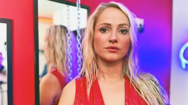 Portrait of Mel, who is looking at the camera with a serious expression on her face. She has long, blonde, wavy hair and is wearing a red, v-necked dress which is patent red in colour. She stands in front of a mirror, with a chain hanging from the ceiling and neon lights also in shot. 