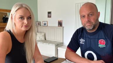 Emma and James Richardson sit at a table in their smartly decorated home. Emma has long blond hair and is wearing a black sleeveless top. James has short fair hair and a neatly trimmed beard and is wearing a blue England rugby shirt. 