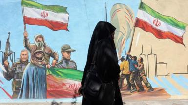 woman in chador walks past wall painted with people holding Iranian flags