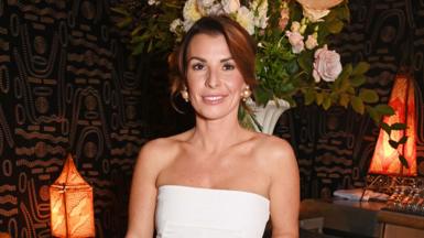 Coleen Rooney wearing a white dress standing in front of a bunch of flowers in a room with black and gold patterned wallpaper