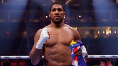Anthony Joshua raising a fist with a smile at the camera while stood in a boxing ring