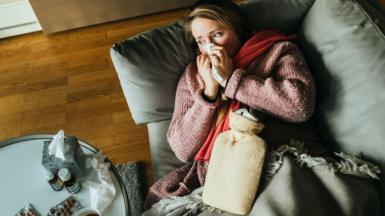 A woman lying on a sofa while having a flu and feeling sick, with a hot water bottle on her stomach and medicine on the table