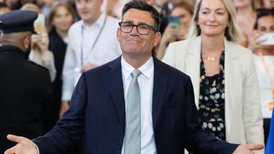 Andy Burnham, wearing a dark suit, silver tie and white, with arms open and a crowd behind him