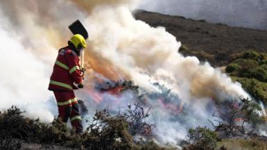 A fireman attends a wildfire in North Wales. Smoke can be seen billowing from a burning moor and a fireman with a shovel is in the foreround