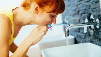 Woman with red hair leans over a sink with the tap running to clean her teeth