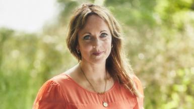 Jenny Evans smiles at the camera. She is wearing a coral pink dress and a necklace. Her blonde hair is pulled to the side and is resting on her shoulder. Behind her is a blurred background of a garden.