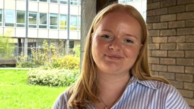 A head-and-shoulders shot of Isobel, outside her office at work, smiling into the camera. She has long, strawberry-blonde hair and wears a striped blue and white shirt.