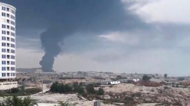 Smoke rises in the distance over a view of a desert-like plane against a cloudy grey sky with buildings in the foreground, in a grab from video showing a strike on Qeshm on 30 July