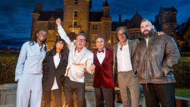Cat Burns, Claudia Winkleman, Alan Carr, Nick Mohammed, David Olosoga and Joe Marler standing outside the Traitors castle at night. Alan Carr is waving one arm in the air in victory.