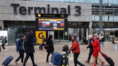 Passengers push luggage outside Terminal 3 as a gate information screen can be seen in the background
