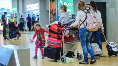 A family with two small children push a luggage trolley pilled with red and pink suitcases through an airport . One of the chidlren is sitting on top of a suitcase and being pushed through the airport by a parent