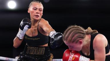 Tiah-Mai in the ring, she black boxing gloves on, black short and top, her is braided. The other boxer is wearing red gloves and is crouching down in the right hand side. 
