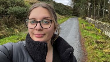 Weronika takes a selfie on a country lane, with a path leading off behind her and hedges on her right side, a row of trees on her left. It's a damp day and she is wearing a black puffer jacket with a fleece lining. She is also wearing stylish large-framed glasses, her light brown hair pulled back in a pony tail.