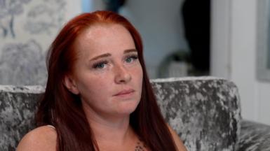 A woman with red hair and blue eyes looks at the camera. She's sitting on a grey velvet-style sofa.