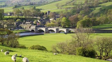 A low stone bridge spans a rural village. It is is surrounded by verdant fields rising up on all sides.