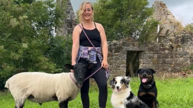 Robyn with a sheep and two dogs in front of a ruined building