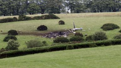 Scene of a helicopter crash in a field. Debris from the grey military helicopter is strewn across the grass. Wreckage can be seen across parts of the field.