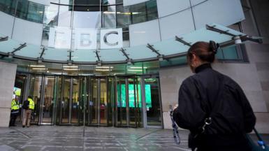 A person wearing a black jacket and carrying a backpack walks towards the entrance doors at BBC Broadcasting House in London on Monday, with the BBC logo in view.