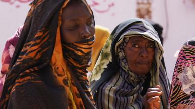 Women in colourful headscarves wait in a queue for food to be distributed.