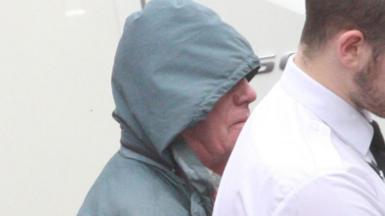 Alan MacPherson is wearing a blue waterproof jacket with the hood up as he is led away by a custody officer 