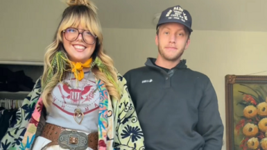 Jessica Raialo wearing a green and blue flower patterned jacket, orange neck scarf, grey and red t-shirt and a belt, next to her boyfriend wearing a dark cap and long-sleeved top.
