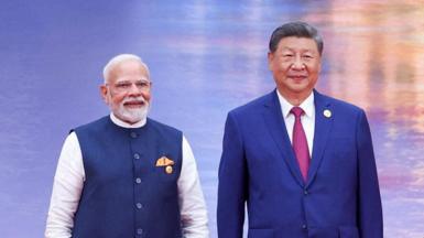 Narendra Modi and Xi Jinping smile for the cameras on stage against a shimmering blue and orange backdrop. Modi is wearing a blue vest over a white kurta with a golden pocket handkerchief, Xi is wearing a navy blue suit and a maroon tie.
