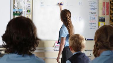 A girl in a light bue shirt and tie writes on a whiteboard. Other students are looking at the board. 