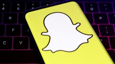 A closeup of Snapchat's logo, a white cartoon ghost with a bright yellow background, held over a laptop conputer keyboard.