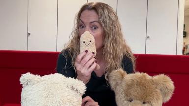 Women with blonde, wavy hair is sitting on a red sofa with teddy bears in front of people.