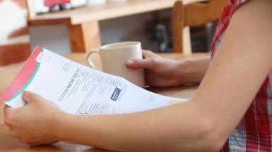 A woman wearing a checked top sits at a wooden table, holding a bill in one hand and a mug in the other.