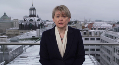 Foreign Secretary Yvette Cooper speaking from a conference in Munich. Shge wears a black overcoat with a white scarf and a blue beaded necklace. She has short, ash blonde hair with a fringe and two silver stud earrings. Snow-covered buildings can be seen behind her rooftop position. Copyright