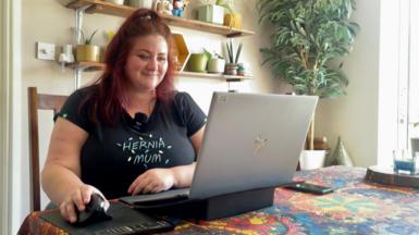A woman sits scrolling a mouse at a laptop in a dining room. There are plant pots behind her on two shelves.